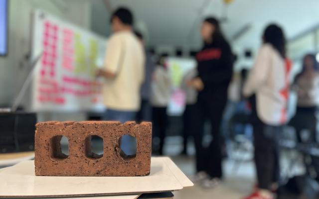 A brick in the foreground and students standing in front of white boards filled with colorful sticky notes in the background